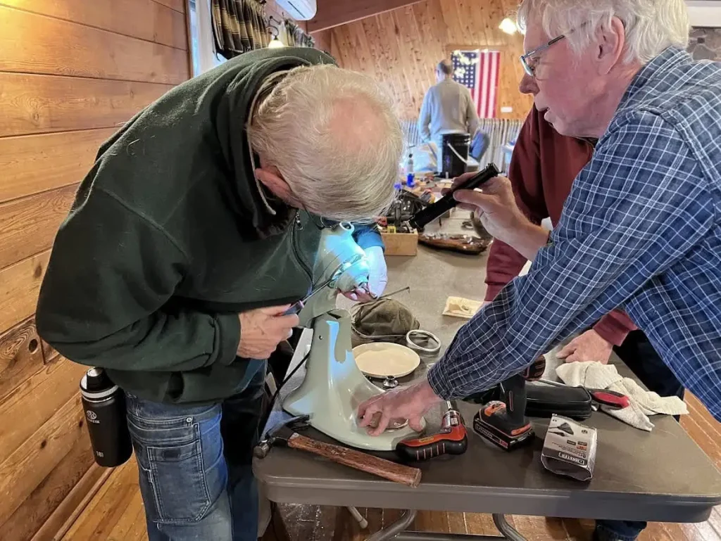 Three men hovered around a small appliance on a table top, working together to fix it.