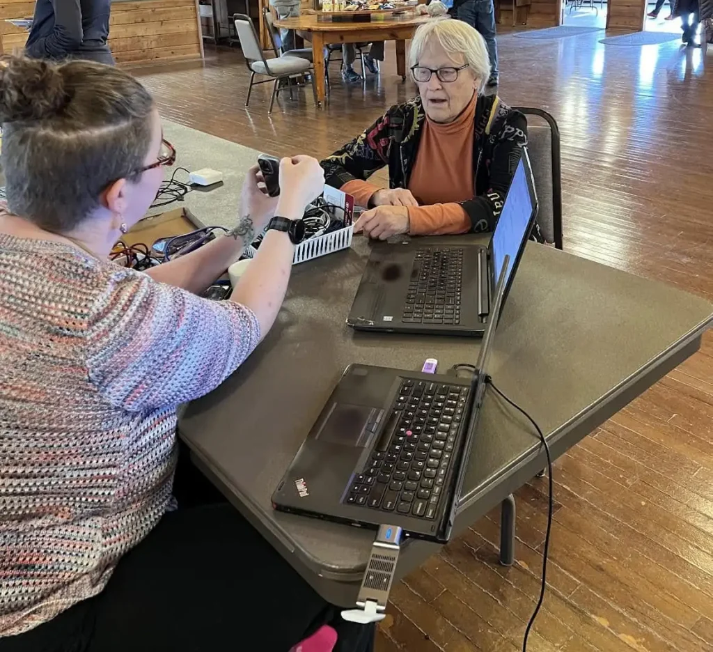 Two women sitting across from each other at a table with a variety of computer equipment displayed on the tabletop.
