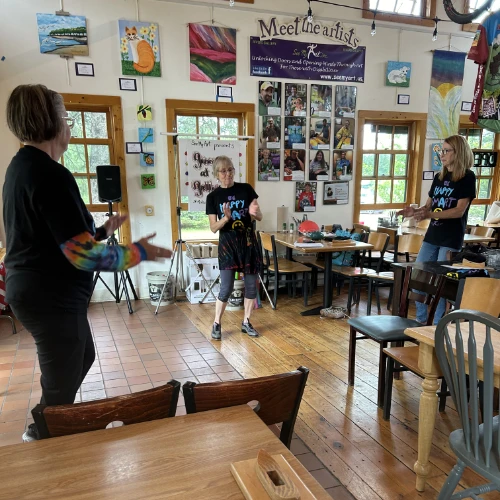 Three women stand in a brightly lit art studio, leading a music and movement activity during Make Music Day. They wear matching “Happy Craft” shirts and appear mid-conversation or demonstration. Colorful artwork and artist bios fill the walls behind them, creating an inviting and energetic space.