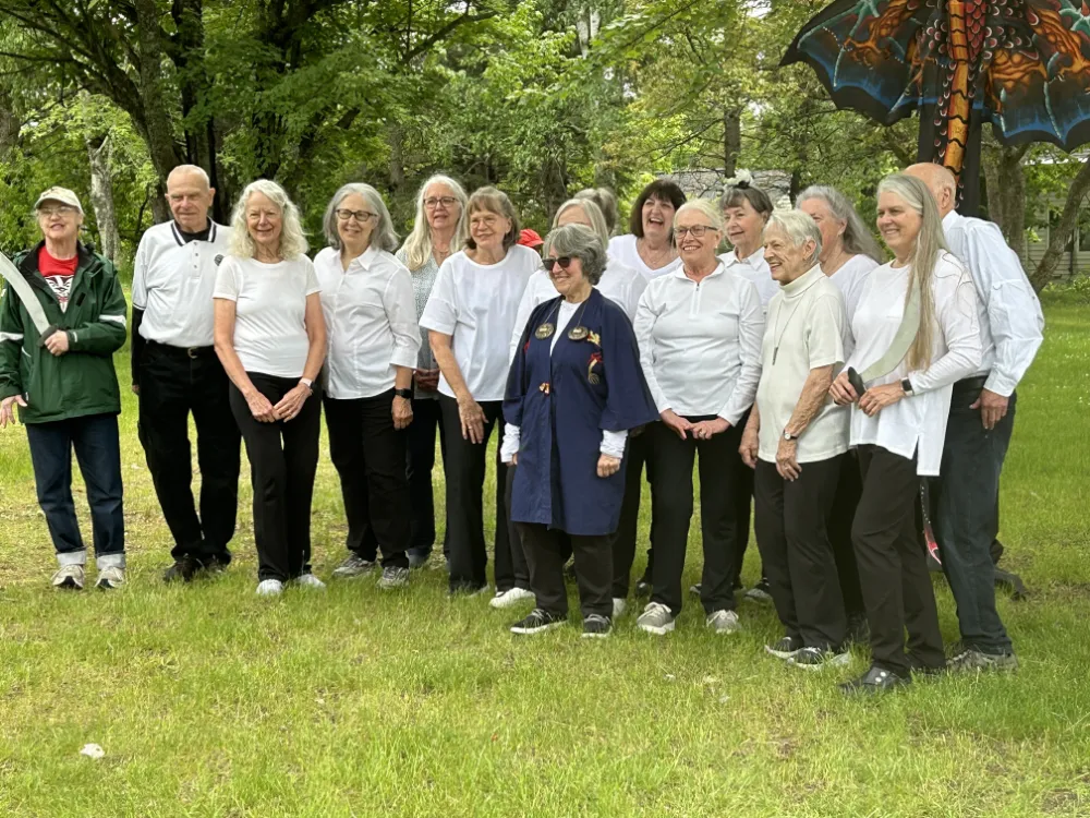 A group of older adults pose together outdoors on a grassy lawn, smiling and laughing under the trees. Most are wearing black pants and white tops, gathered for a group photo during Make Music Day. A colorful dragon kite peeks in from the upper right corner.