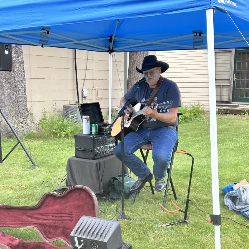 A man in a cowboy hat sits under a blue canopy playing an acoustic guitar and singing into a microphone. He’s performing outdoors on a grassy lawn with audio equipment nearby and a guitar case open in the foreground.